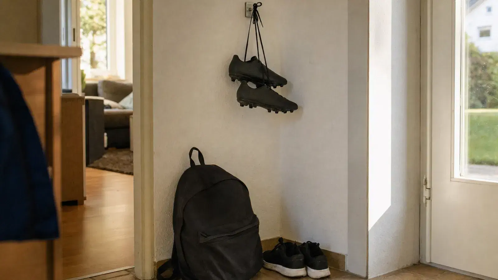 Ein Paar schwarze Fußballschuhe hängt an den Schnürsenkeln an einem Garderobenhaken im Hausflur, darunter stehen ein Schulrucksack und ein Paar Turnschuhe.