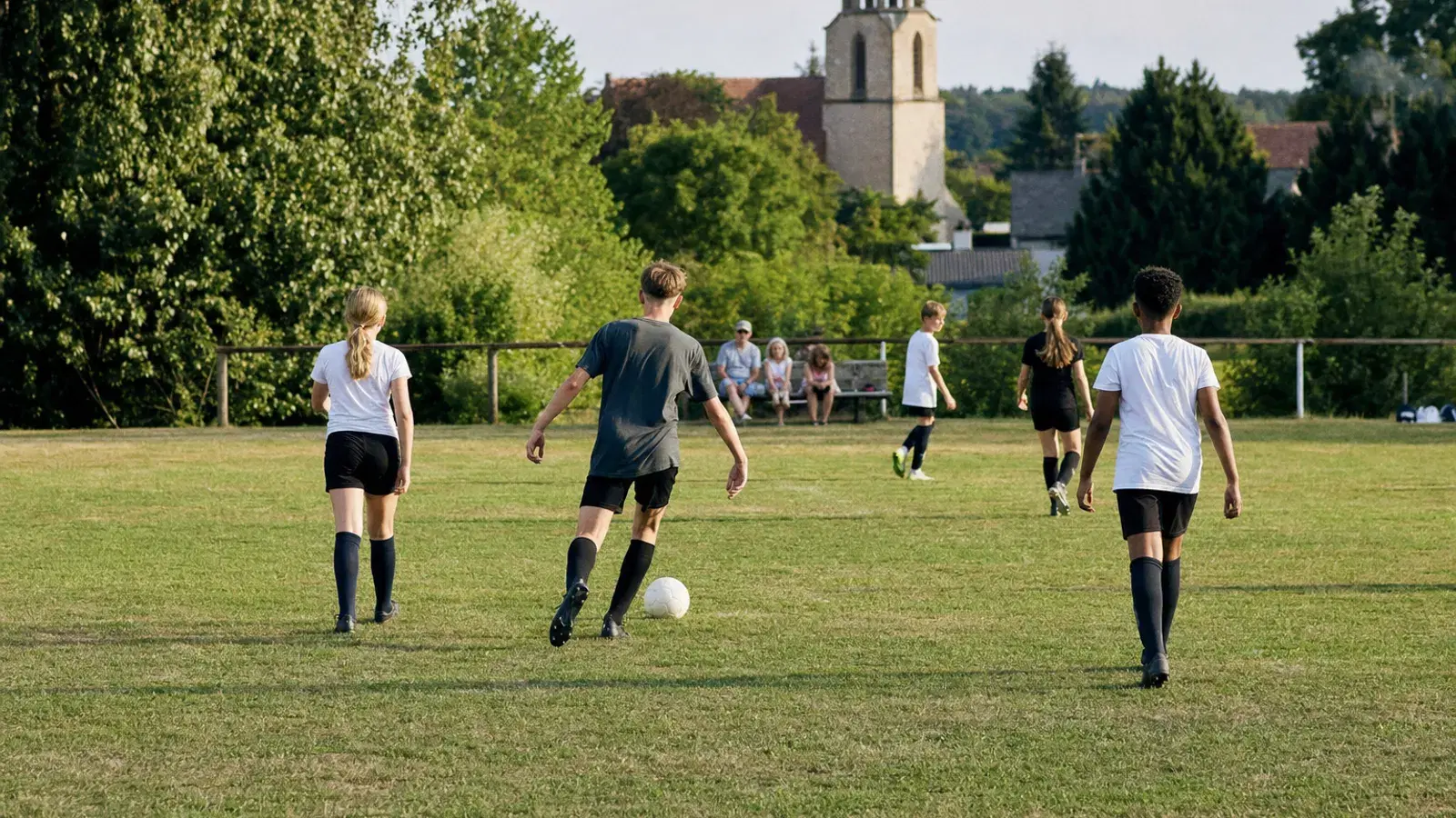 Mehrere Jugendliche spielen an einem sonnigen Nachmittag entspannt Fußball auf einem Dorfplatz, aus der Distanz gesehen.