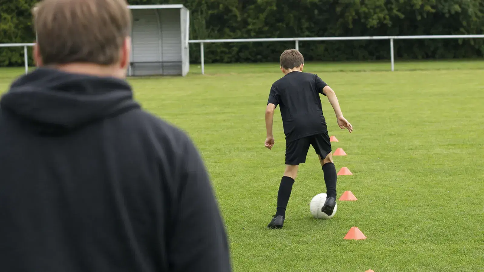 Ein etwa neunjähriges Kind dribbelt von hinten gesehen einen Ball durch einen Hütchenparcours, im unscharfen Vordergrund steht ein Elternteil.