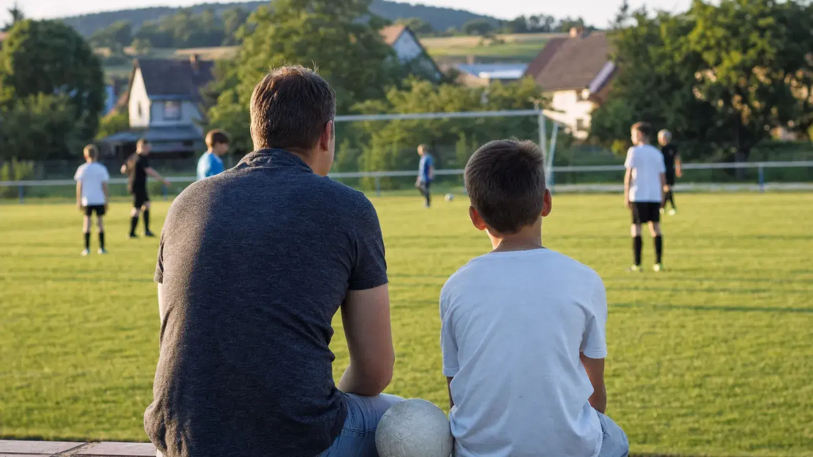 Ein Vater und sein Sohn sitzen von hinten gesehen nebeneinander auf einer niedrigen Mauer und schauen auf einen Fußballplatz.