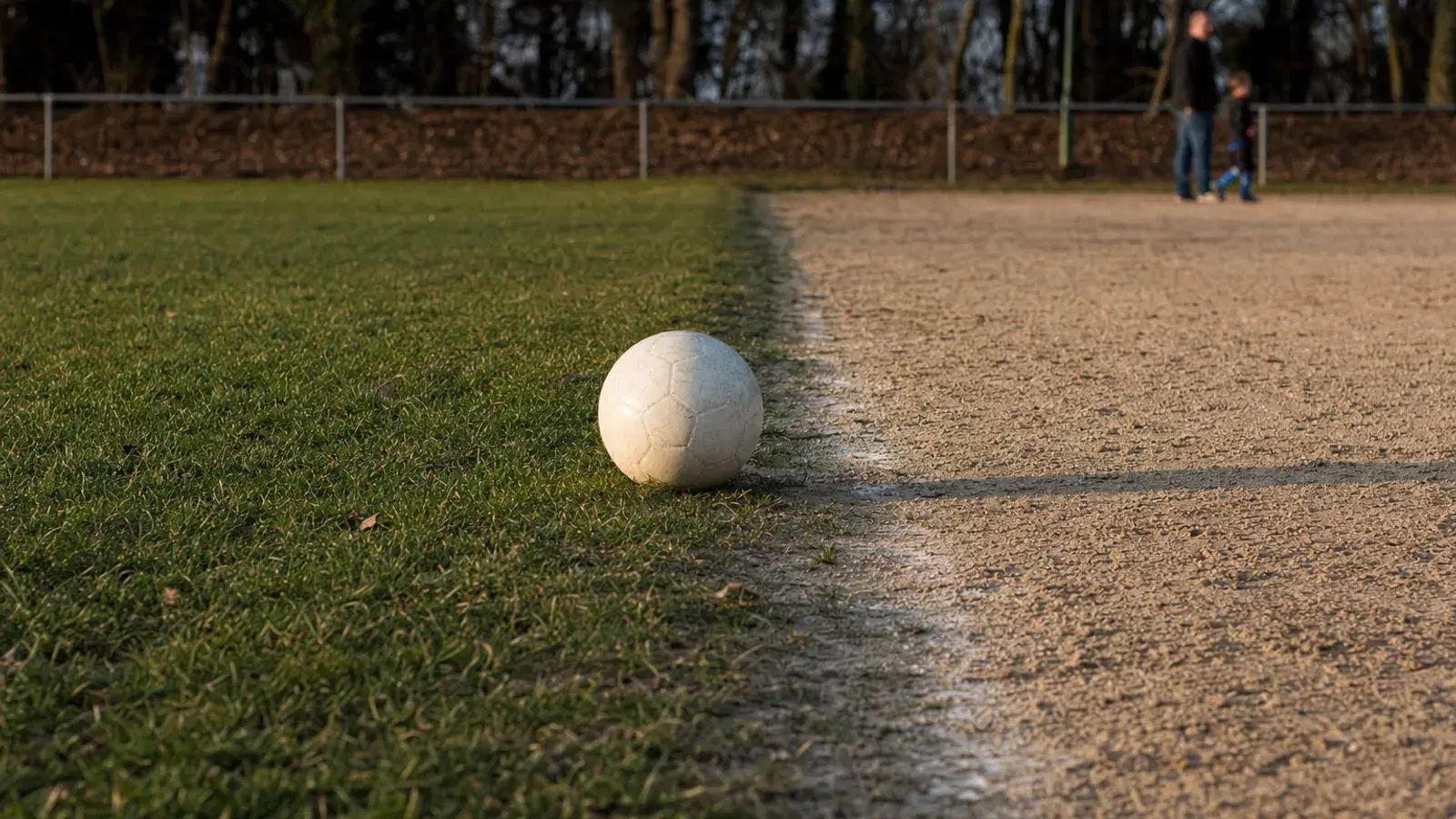 Ein abgenutzter weißer Fußball liegt genau an der Kante, an der ein Rasenplatz in einen sandigen Hartplatz übergeht, im flachen Abendlicht; weit hinten gehen unscharf ein Erwachsener und ein Kind am Zaun entlang.