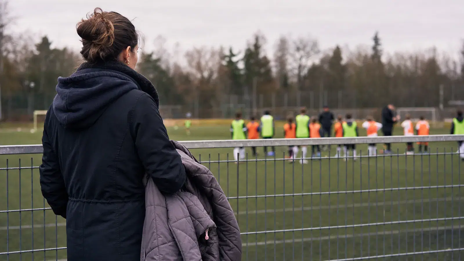 Eine Mutter steht von hinten gesehen am Zaun eines Trainingsplatzes und hält eine Kinderjacke über dem Arm, in der Ferne trainieren Kinder.