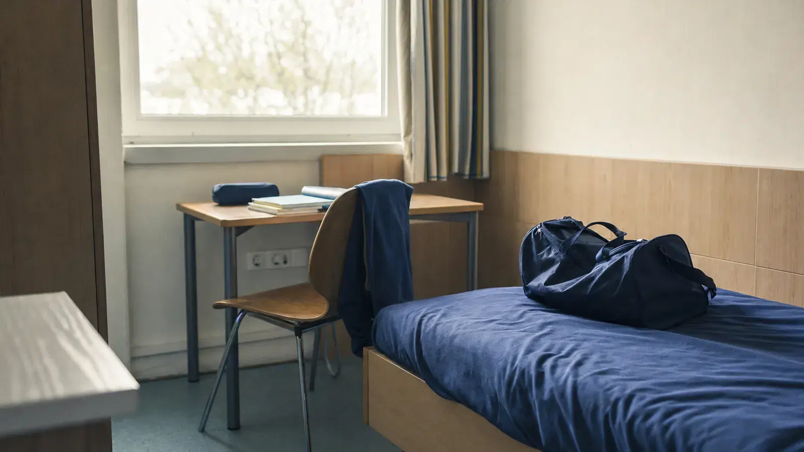 Ein schlichtes Internatszimmer mit Schreibtisch, geschlossenen Schulbüchern, einer gefalteten Trainingsjacke und einer Sporttasche auf dem Bett.