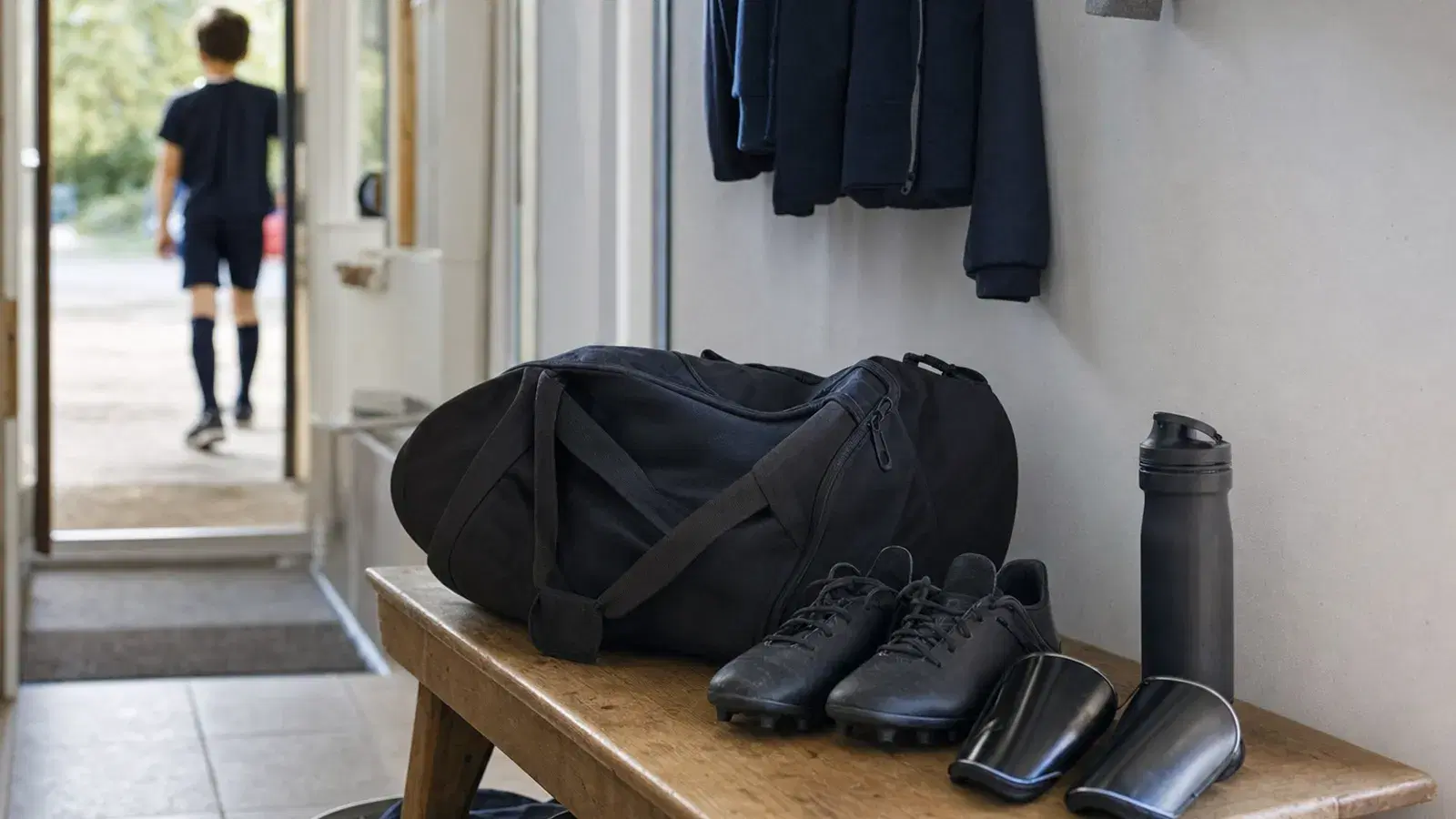 Eine gepackte Sporttasche steht auf einer Bank im Hausflur, daneben Fußballschuhe, Schienbeinschoner und eine Trinkflasche.