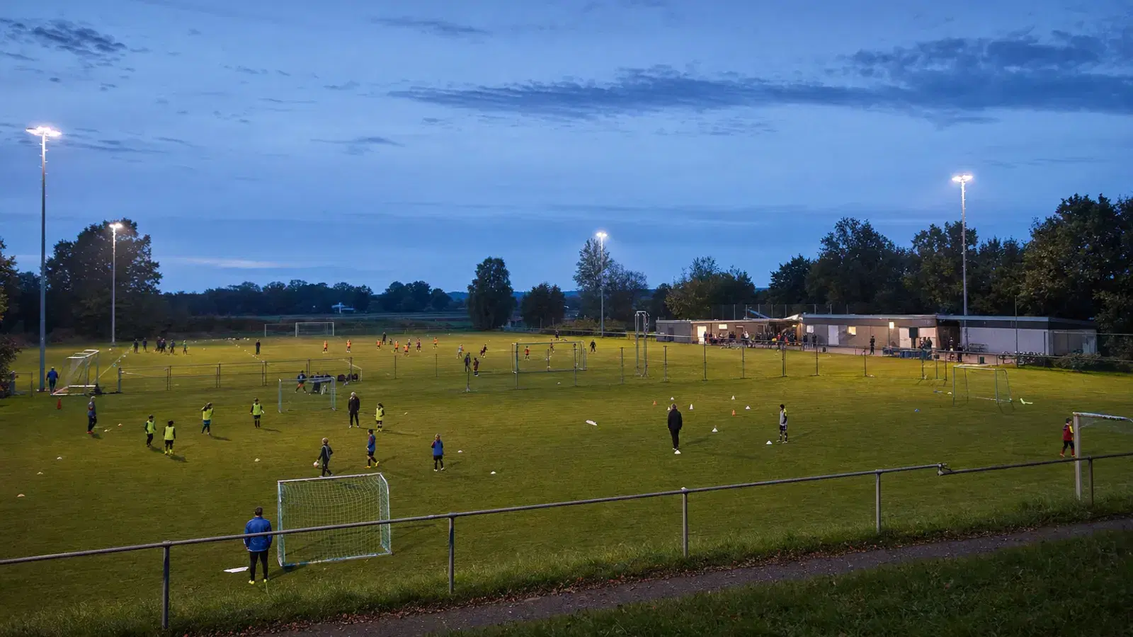 Mehrere Rasenplätze am frühen Abend aus der Distanz gesehen, unter Flutlicht trainieren kleine Gruppen von Kindern und Jugendlichen.