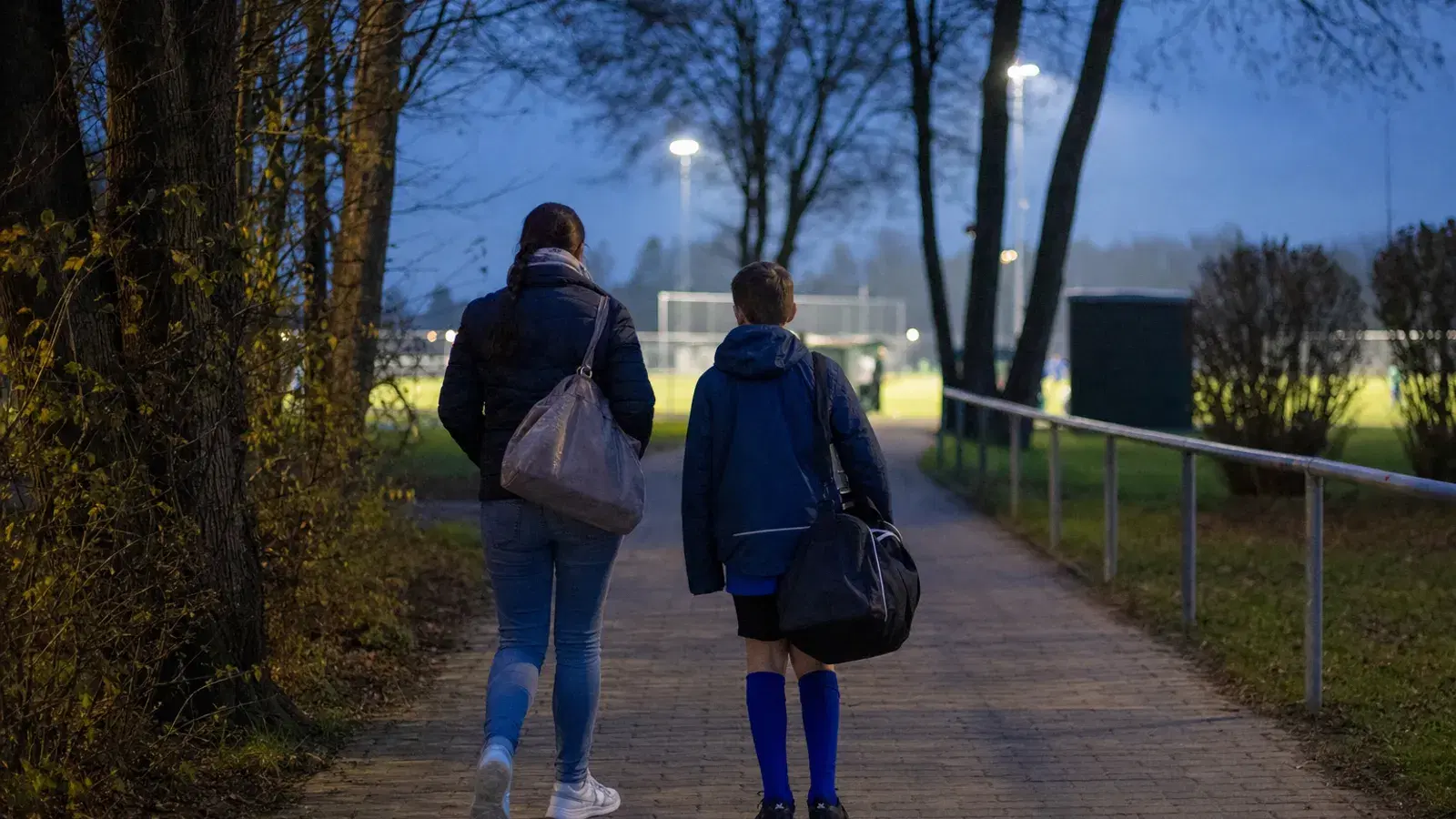 Eine Mutter und ihr Kind gehen am frühen Abend von hinten gesehen einen Weg entlang auf eine beleuchtete Trainingsanlage zu.