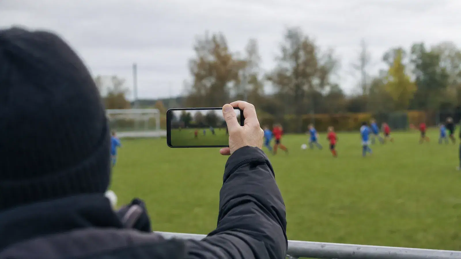 Eine erwachsene Hand hält am Spielfeldrand ein Smartphone hoch und filmt ein Jugendspiel, die Kinder auf dem Platz sind klein und unscharf.