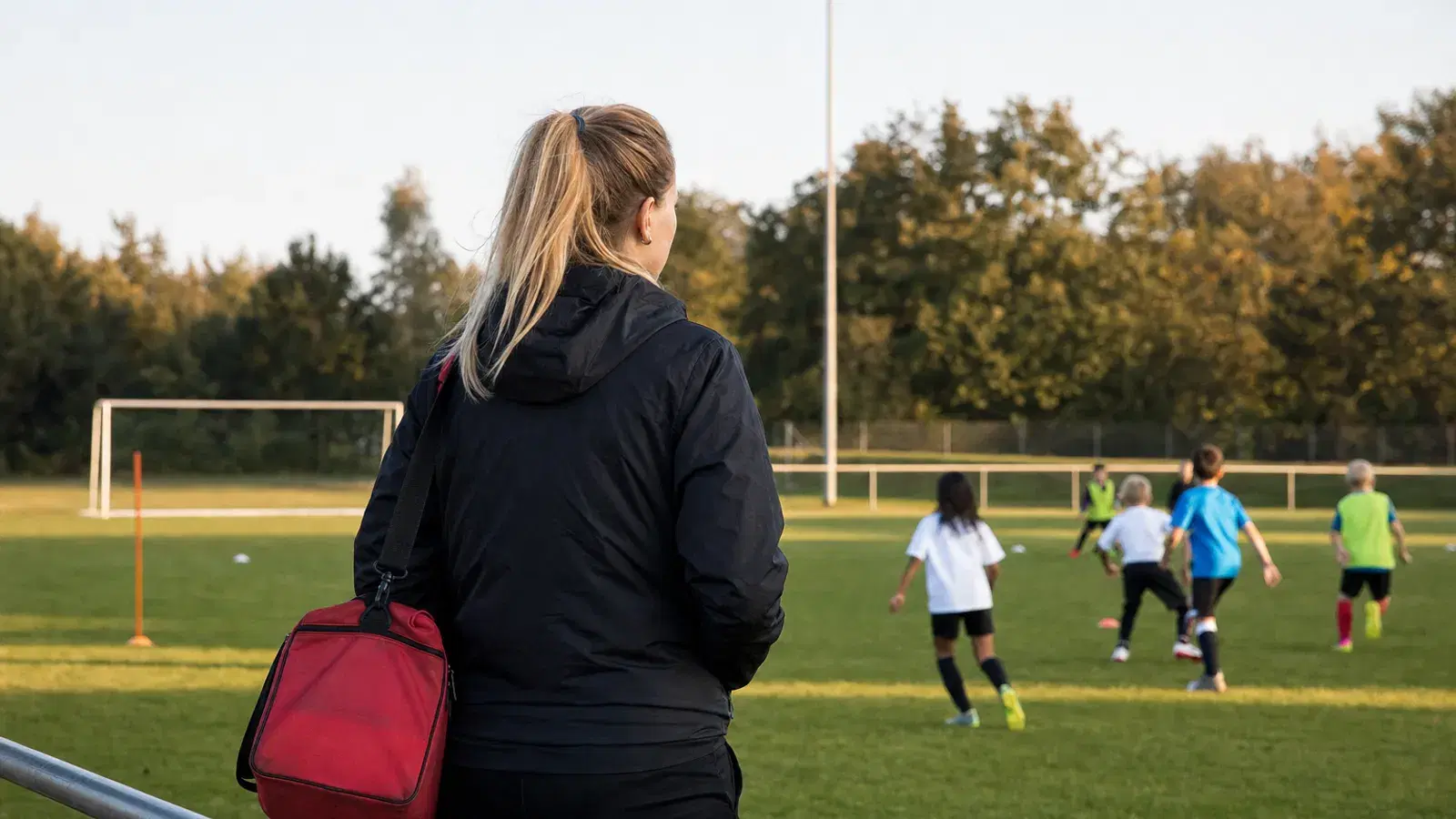 Eine Betreuerin mit einer roten Erste-Hilfe-Tasche steht am Rand eines Trainingsplatzes und beobachtet trainierende Kinder.