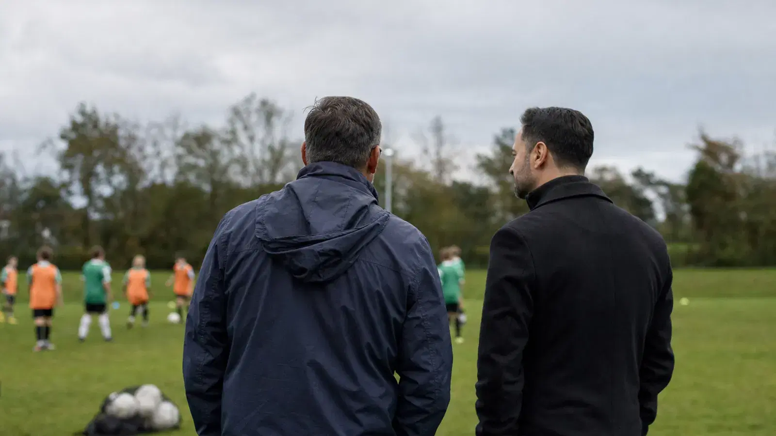 Ein Vater und ein Mann in einer Jacke unterhalten sich von hinten gesehen am Spielfeldrand, in der Ferne trainieren Jugendliche.