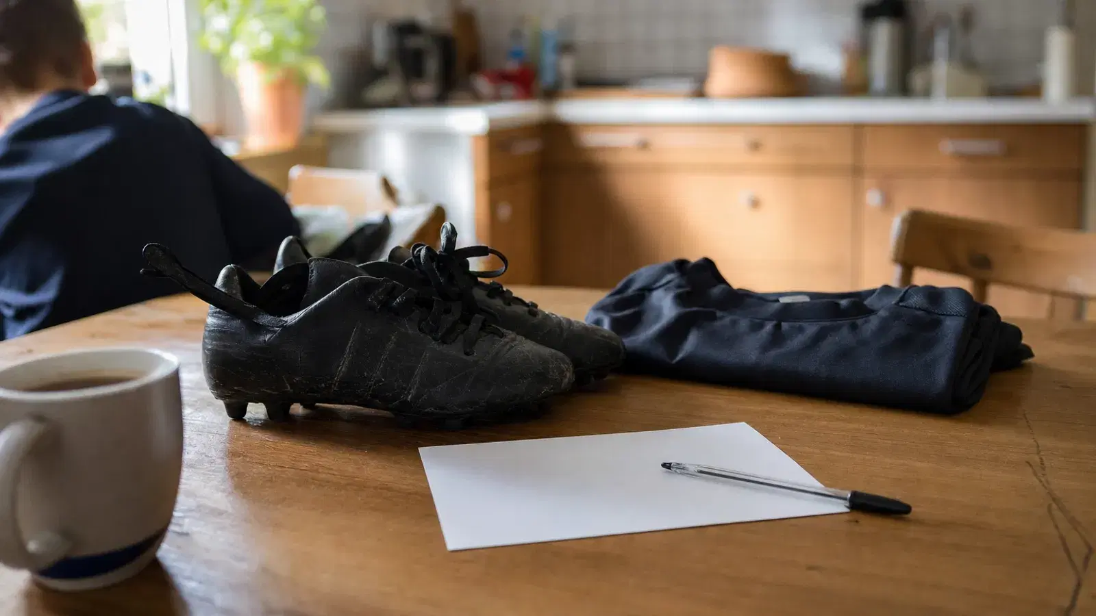 Auf einem Holztisch liegen eine unbeschriebene Postkarte und ein Kugelschreiber, daneben ein Paar schwarze Kinder-Fußballschuhe und ein gefaltetes Trainingsoberteil; am linken Bildrand sitzt unscharf eine Person von hinten gesehen, vorne steht eine Tasse.