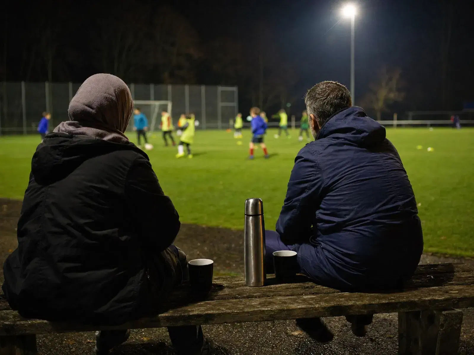 Zwei Eltern sitzen am Abend auf einer Holzbank am Rand eines Trainingsplatzes, eine Thermoskanne steht zwischen ihnen, im Hintergrund trainieren Kinder.