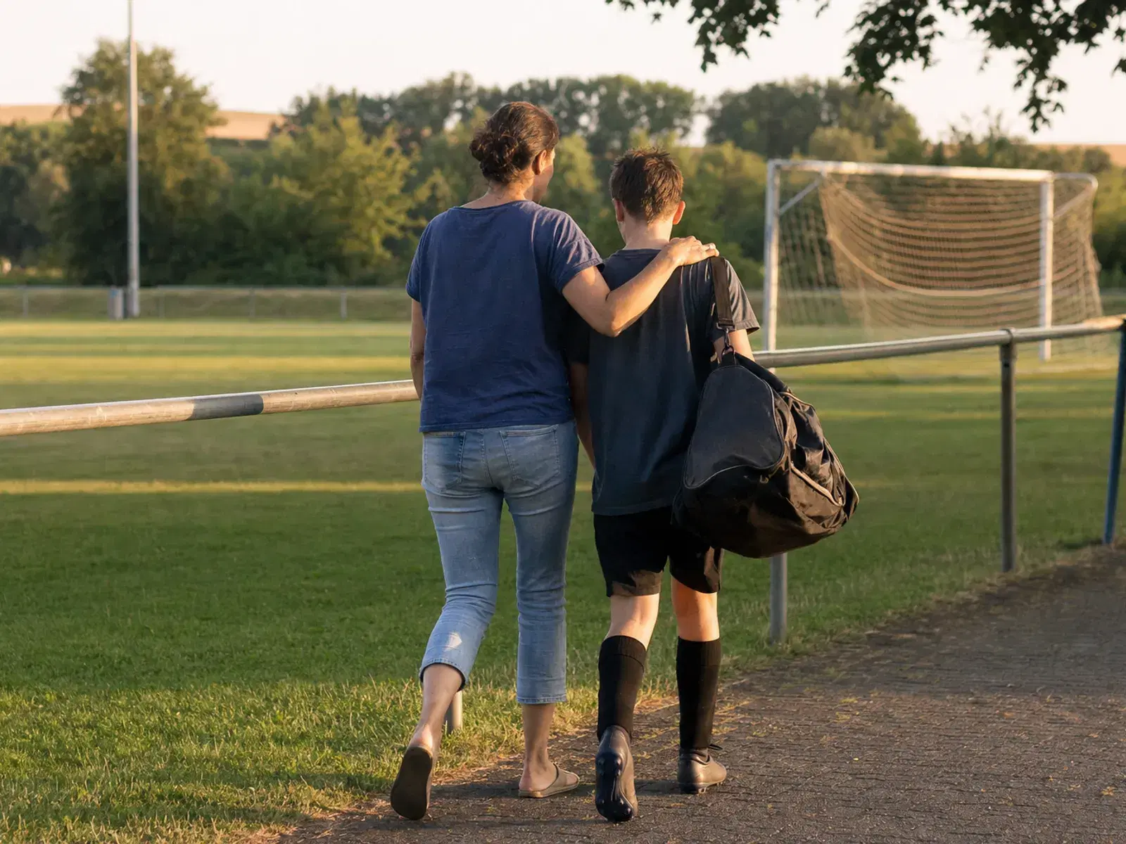 Eine Mutter und ihr jugendlicher Sohn gehen im Abendlicht von hinten gesehen nebeneinander an einem leeren Fußballplatz entlang, ihr Arm liegt auf seiner Schulter.
