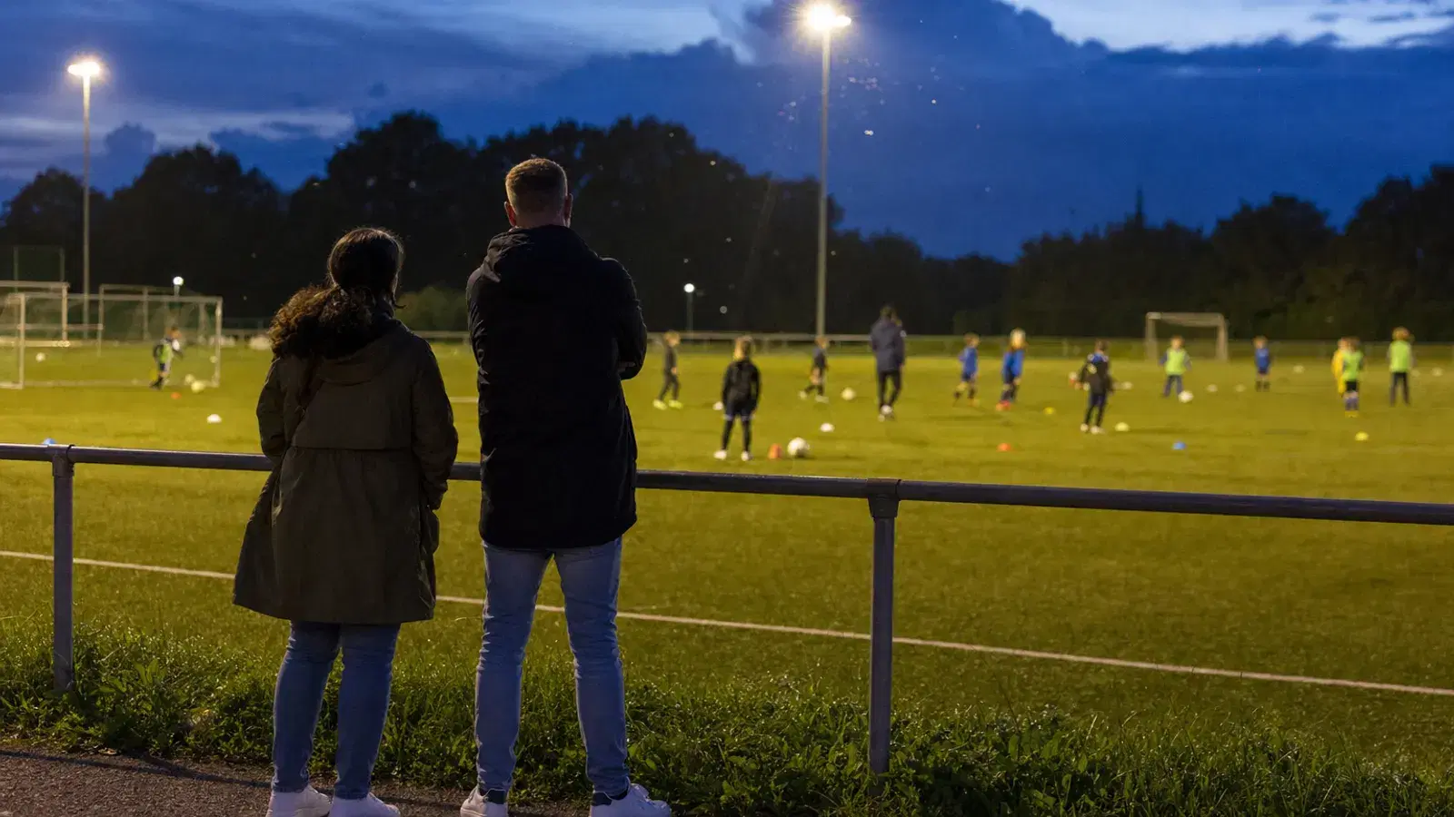 Zwei Eltern stehen am Abend von hinten gesehen am Geländer eines Trainingsplatzes unter Flutlicht, in der Ferne trainieren Kinder.