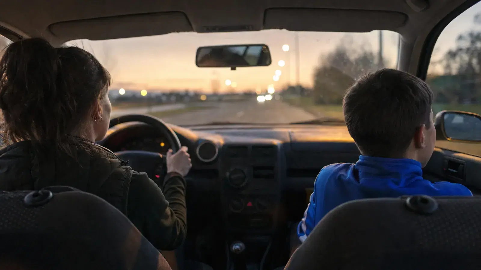 Blick vom Rücksitz eines Autos am Abend: eine Mutter fährt, daneben sitzt ein Kind in Trainingsjacke, draußen leuchten Straßenlaternen.