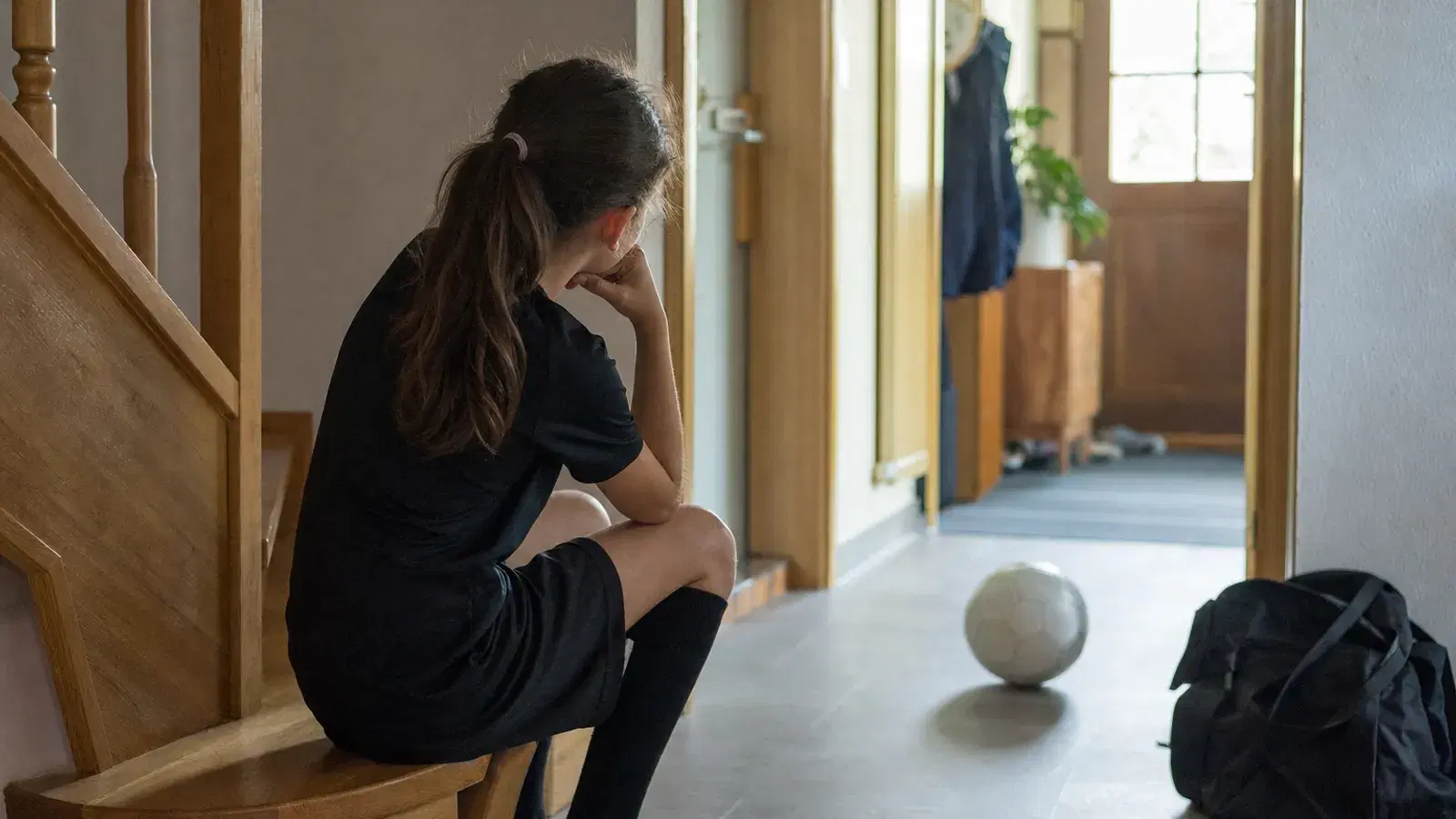 Ein Kind sitzt von hinten gesehen auf einer Treppe im Hausflur, vor ihm liegt ein Fußball unbeachtet auf dem Boden.