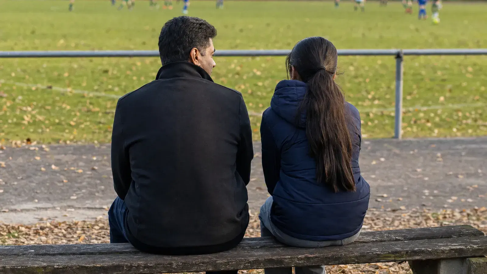 Ein Vater und seine jugendliche Tochter sitzen von hinten gesehen auf einer Parkbank neben einem Fußballplatz und unterhalten sich.