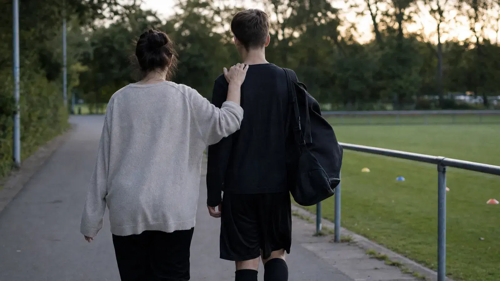 Eine Mutter und ihr jugendlicher Sohn gehen am Abend von hinten gesehen nebeneinander auf einem Weg neben einem leeren Trainingsplatz, sie hat die Hand leicht auf seine Schulter gelegt, er trägt eine Sporttasche.