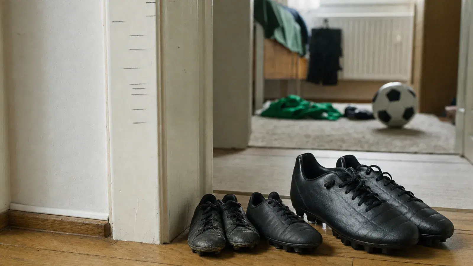 Ein weiß gestrichener Türrahmen im Flur mit mehreren Bleistiftstrichen in verschiedenen Höhen, daneben stehen schwarze Fußballschuhe in mehreren Größen, von klein und abgetragen bis groß und neu; im Hintergrund liegt ein Ball in einem Kinderzimmer.