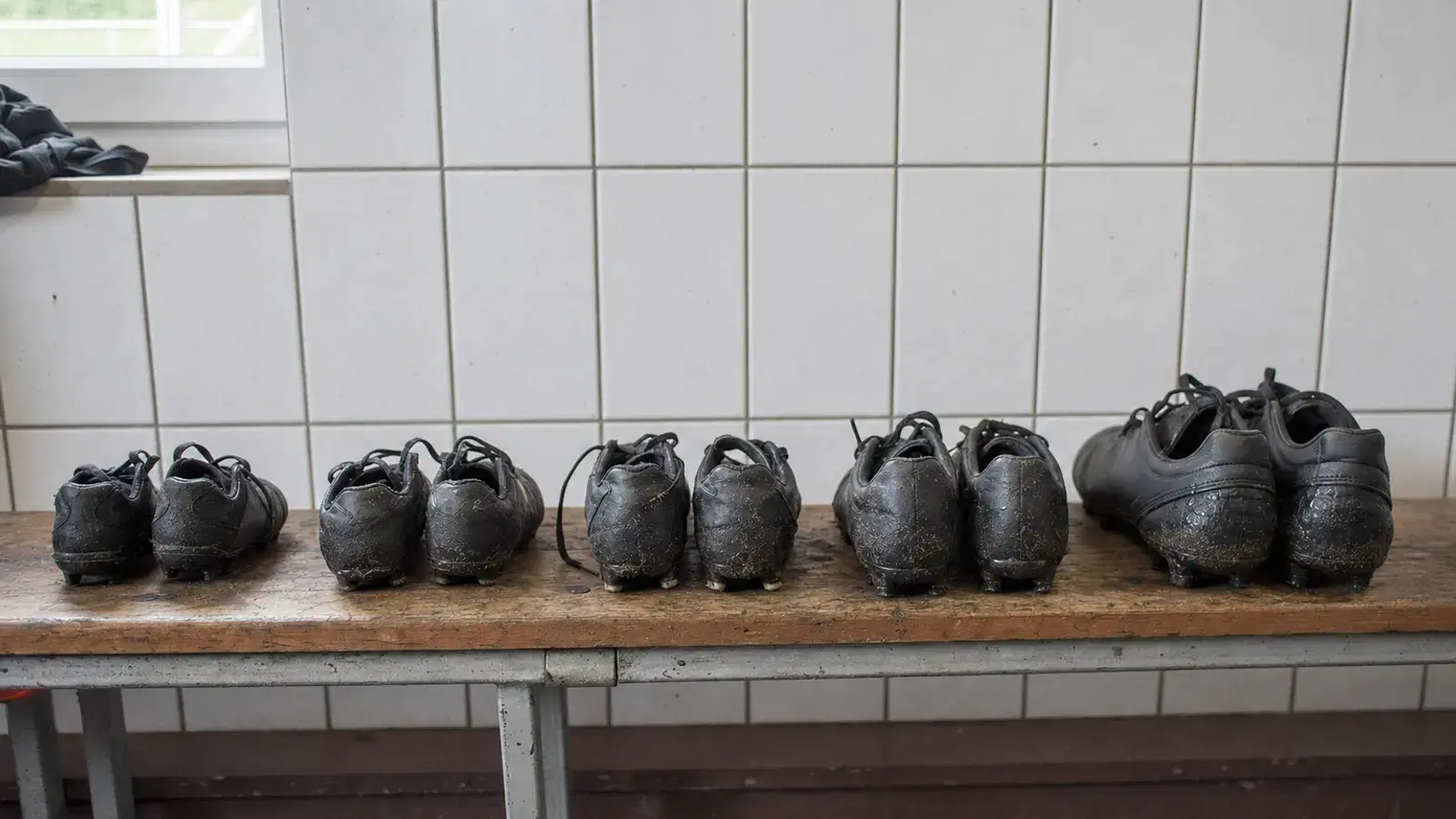 Fünf Paar schwarze Fußballschuhe in aufsteigenden Größen stehen nebeneinander auf einer Holzbank in einer Umkleide.